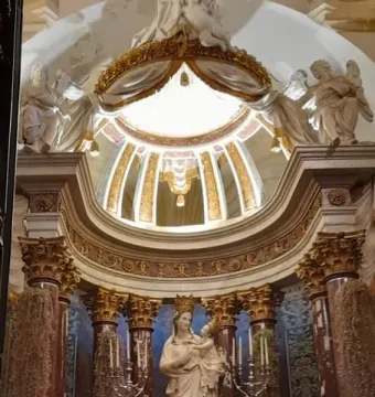 Crowned Madonna di Trapani statue holding the Child, set in an ornate altar with angels and pink flowers.