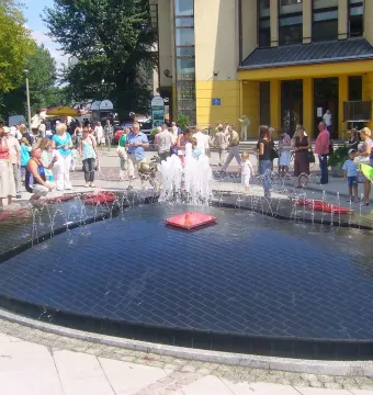 A small bronze Reksio monument beside a large fountain with water jets and people gathered around.