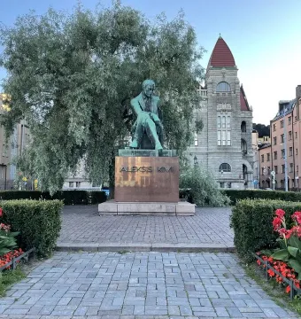 Aleksis Kivi Memorial bronze statue of a seated figure on a pedestal in a paved square, with hedges, red flowers, and buildings behind it.