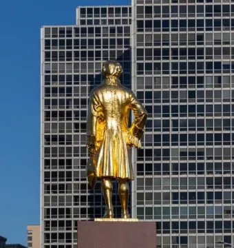 Alexander Hamilton Memorial statue in colonial attire on a stone pedestal against a blue sky and a modern building.