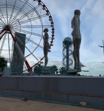 Ali and Nino sculpture with two metallic human figures facing each other on a platform, with a Ferris wheel in the background.