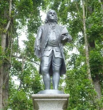 Benjamin Franklin statue in colonial attire on an ornate stone pedestal, framed by trees.