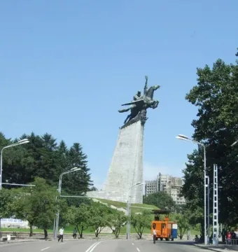 The Chollima Statue rises above a multi-lane road in Pyongyang, with trees, streetlights, and buildings in the background.