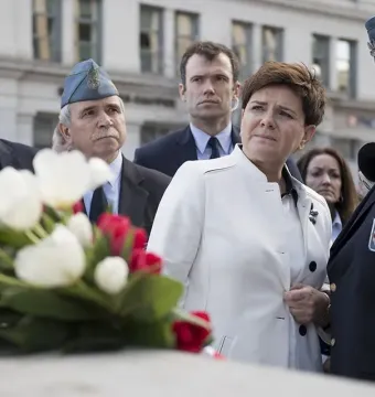 People gathered at the Katyń Memorial with white and red flowers in the foreground and a man in a blue uniform speaking into a microphone.