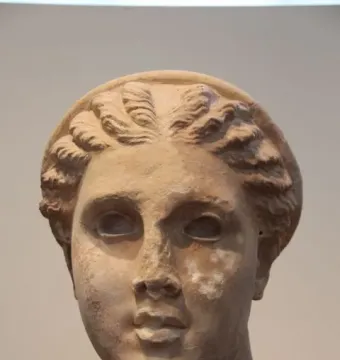 Weathered stone head of Lycosoura Artemis with intricate wavy braids and a headband, mounted on a dark base.