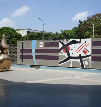 Cloud Shepherd, a golden-bronze abstract sculpture, stands beside a colorful geometric wall in a plaza.