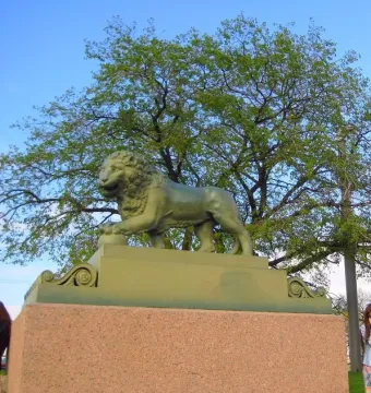 Bronze lion sculpture on a tall granite pedestal with a tree and blue sky behind it.