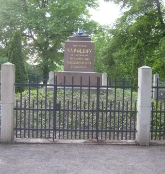 Napoleonstein stone monument behind a black metal fence in Leipzig, framed by green trees.
