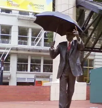 Allow Me bronze sculpture of a suited man holding an open umbrella and raising one hand on a brick plaza.