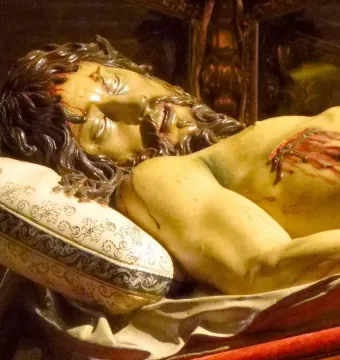 Cristo Yacente wooden sculpture showing the upper body, with dark hair, a beard, and blood on the forehead and chest while resting on an ornate pillow.