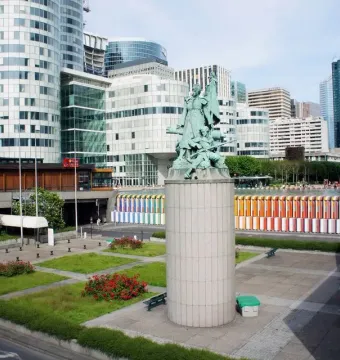 La Défense de Paris sculpture on a tall pedestal in a modern urban plaza with lawns and skyscrapers.