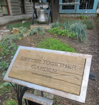 A large silver Liberty Bell display stands in a garden beside a Better Together Garden sign.