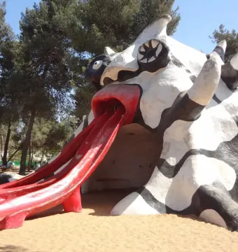 The Golem sculpture with a red mouth and three red slide chutes on sandy ground under a blue sky.