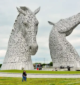 The Kelpies metal horse-head sculptures rising above a grassy field, with visitors near the base for scale.