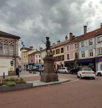 The Statue of the Volunteer of 1792 on a stone pedestal in a cobblestone plaza, with buildings and parked cars nearby.