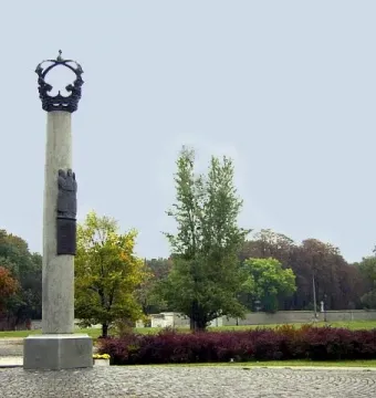 Electio Viritim Monument, a tall pale column topped with a dark crown and relief carvings, standing on a cobblestone base in a park with autumn trees.