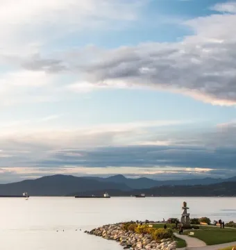 An Inukshuk stands in a grassy park overlooking the water, with distant mountains and ships under a cloudy sky.