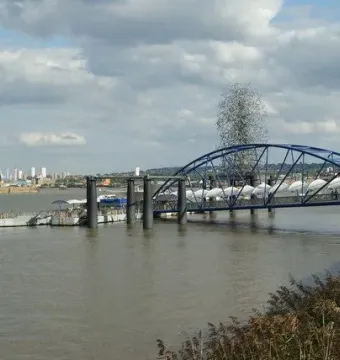 Quantum Cloud sculpture rises behind a blue bridge over the river near a pier.