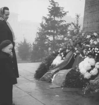 Floral wreaths and ribbons placed beside the Stalin monument, with a woman and child standing nearby.