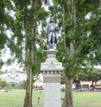 Benjamin Franklin memorial on a stone pedestal in a park, framed by trees and nearby buildings.