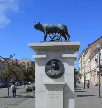 The Capitoline Wolf statue on a tall stone pedestal in a pedestrian plaza, with a circular relief and golden inscription on the base.