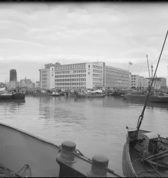 Boats moored in a harbor with multi-story buildings along the far bank under a cloudy sky.