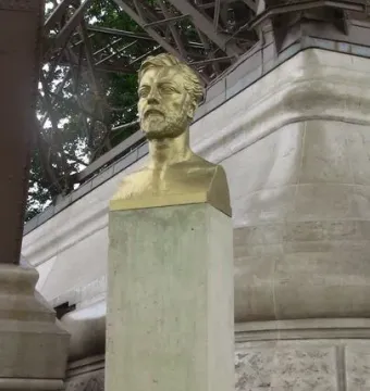 Golden bust of Gustave Eiffel on a stone pedestal with the Eiffel Tower framework in the background.