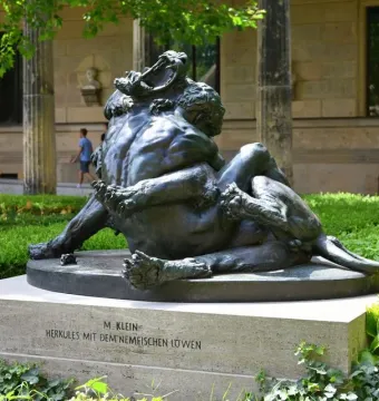 Dark bronze Hercules and the Nemean Lion statue on a stone pedestal beside a grand stone building.