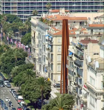 Neuf Lignes Obliques sculpture standing between apartment buildings on a busy city street lined with parked cars and palm trees.