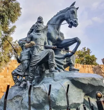 The Statue of Saladin, a bronze equestrian monument with two armed figures beside it, set against stone walls and trees.