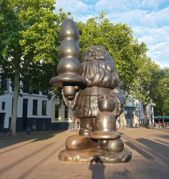 Bronze Santa Claus statue in a plaza, holding an abstract object in one hand and a bell-shaped object in the other.