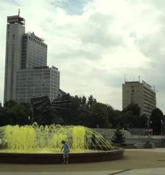 Silesian Insurgents\' Monument partly visible behind a fountain with yellow water jets and trees.