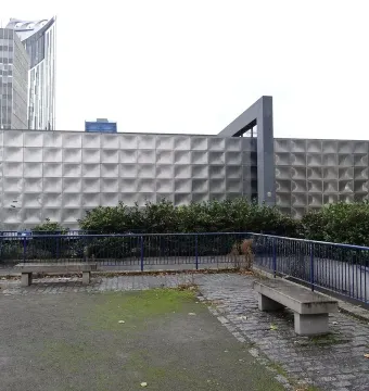 The Michael Faraday Memorial seen behind green bushes and a blue fence, with tall buildings in the background.