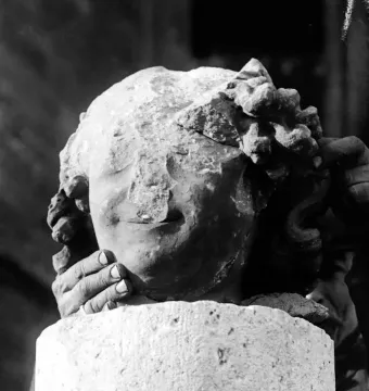 Damaged stone head of the Smiling Angel with a faint smile, held by two hands above a stone pedestal.