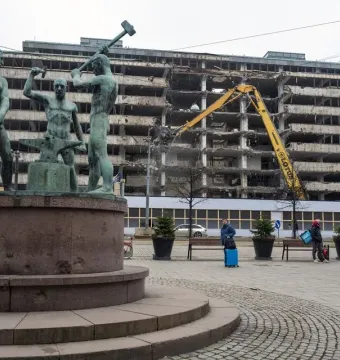 The Three Smiths bronze sculpture stands in front of a building demolition site with a yellow excavator in the background.
