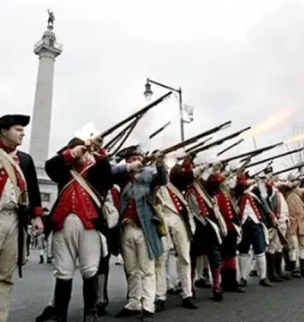 The Trenton Battle Monument rises above colonial reenactors firing muskets in the foreground.