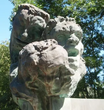 Bronze sculpture on the War Memorial of Capoulet-et-Junac showing three textured human heads against trees and blue sky.