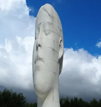 The Dream sculpture by Jaume Plensa, a tall white head and neck with closed eyes, against a blue sky with clouds.