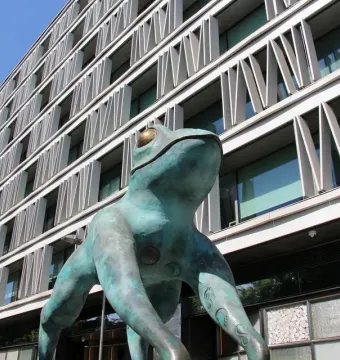 Lucky Frog statue with teal patina and golden eyes in front of a modern building.