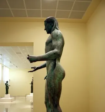 The Piraeus Apollo, a bronze nude male statue with braided hair, shown in profile on a stone base.