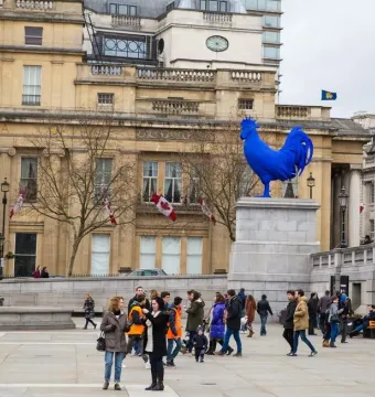 Blue Bird of Canada sculpture on a stone plinth in an urban plaza with buildings and pedestrians behind it.