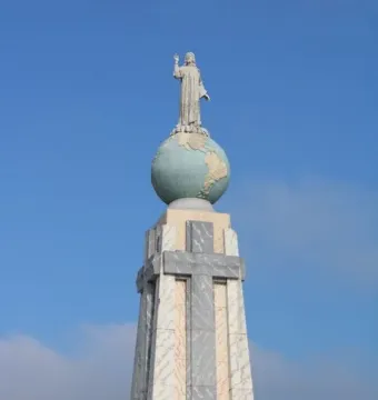 Divino Salvador del Mundo monument showing a robed figure raising one arm atop a globe and stone pedestal against a blue sky.
