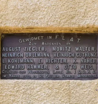 A bronze plaque with raised German text set into a light beige wall at the Hermann Heights Monument.