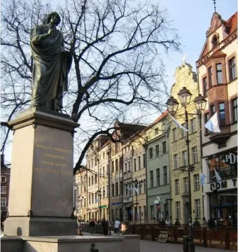 Nicolaus Copernicus Monument on a tall stone pedestal in a historic town square, with a fountain, benches, and old buildings nearby.