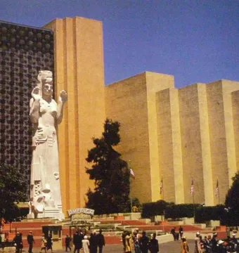 The Pacifica statue stands in a plaza, with raised arms, an elaborate headpiece, and people gathered at its base.