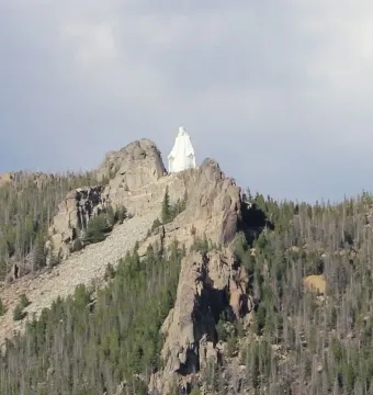 Our Lady of the Rockies statue atop a rugged mountain peak, surrounded by forested slopes under a cloudy sky.