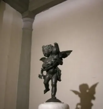 Bronze Putto with Dolphin sculpture of a winged child holding a dolphin on a pedestal, with its shadow cast on the wall.