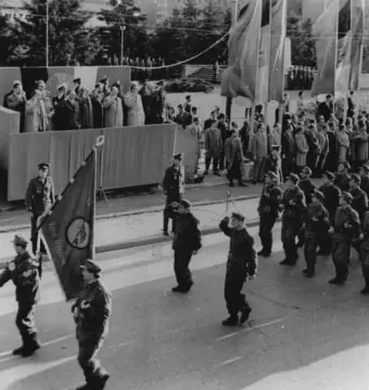A statue of Joseph Stalin stands near a ceremonial parade with uniformed marchers, flags, and spectators on a raised platform.