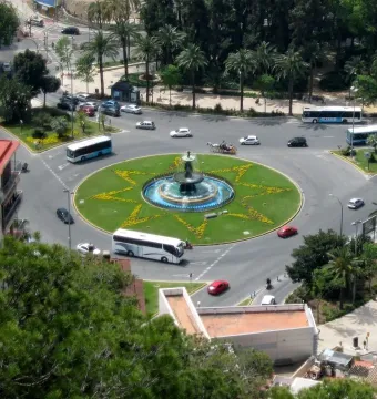 Three Graces Fountain in a traffic roundabout with water flowing into a blue basin and a star-shaped flowerbed around it.