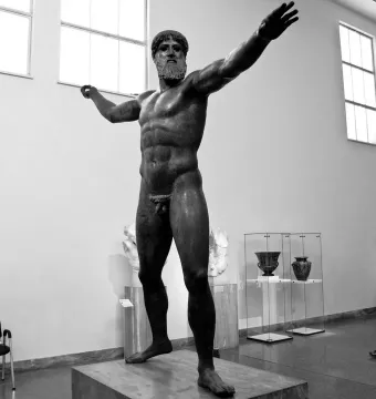 The Artemision Bronze stands on a plinth in a museum hall with arms extended.
